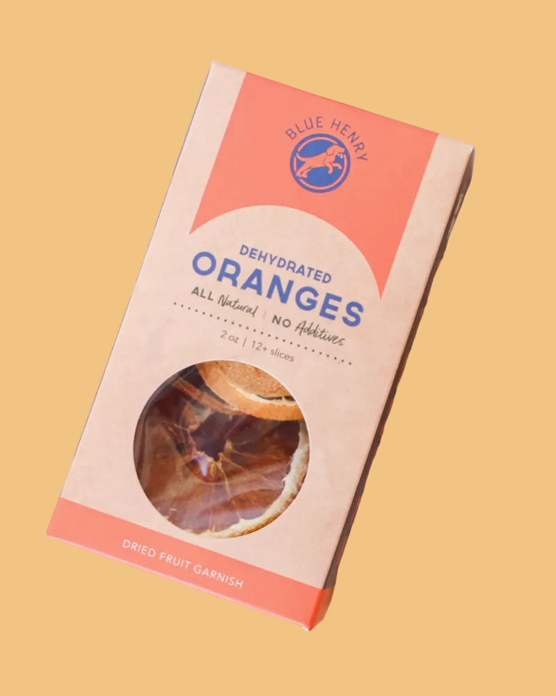 A package of Blue Henry Dehydrated Oranges garnish slices, shown in a styled flat-lay arrangement of dried orange slices on a neutral background.