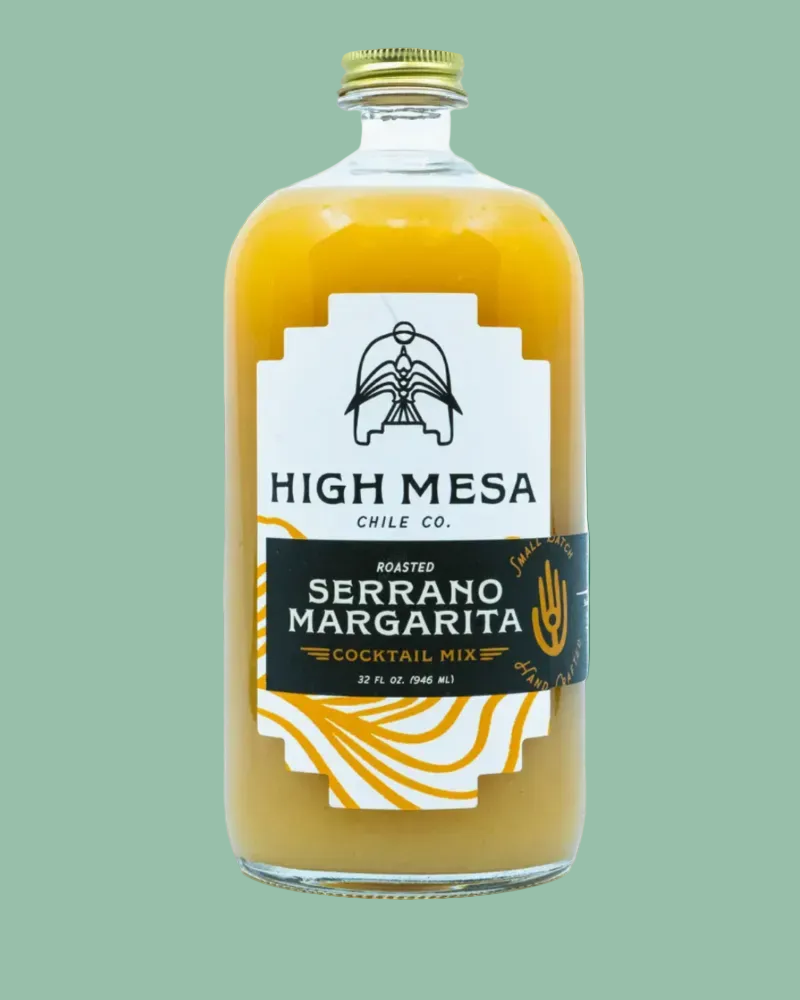 A clear glass bottle of High Mesa Chile Co. Roasted Serrano Margarita cocktail and mix, filled with an orange-yellow liquid, featuring a white and black label with gold accents and a gold cap, on a white background.