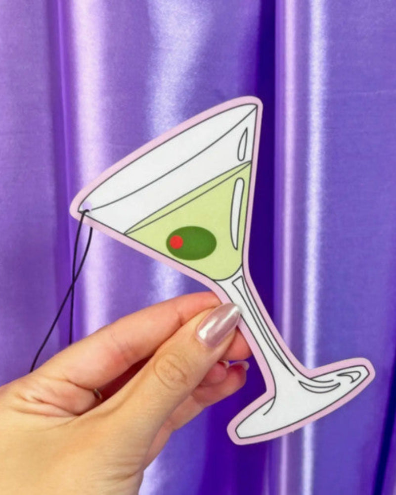 Hand holding a paper cutout of a martini glass with a green cocktail against a purple curtain.
