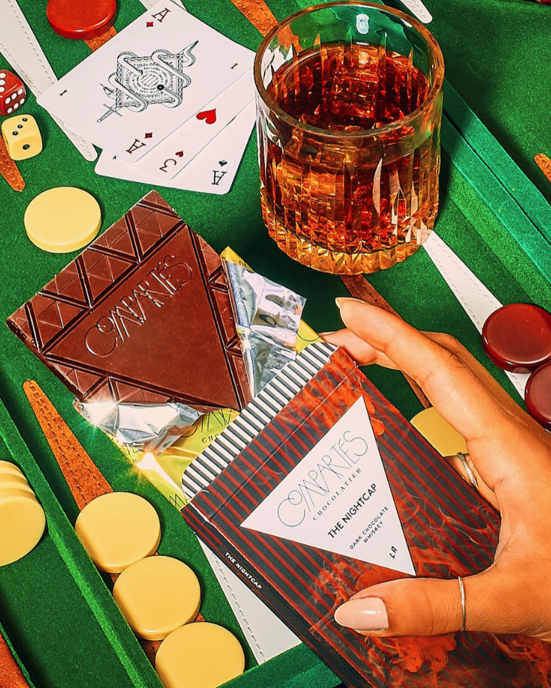Hand holding a Caramba The Nightcap book with a glass of whiskey, chocolate bar, and poker chips on a green table.