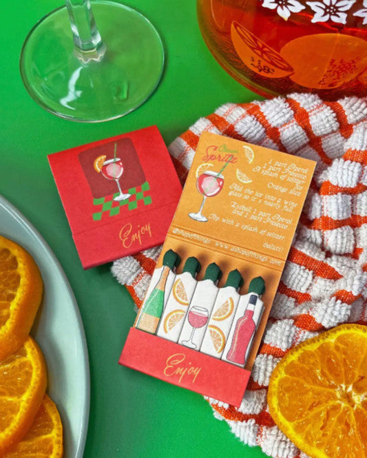 Matches with cocktail design on a green surface with oranges and a glass.