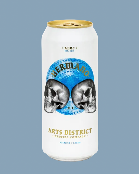 A white aluminum beer can with a gold pull tab and gold text, featuring a blue circular graphic with two black-and-white skulls and the word “Hermano.” The can is centered on a muted blue-gray background.