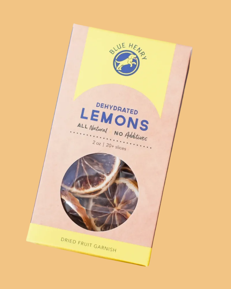 A package of Blue Henry Dehydrated Lemons dried fruit garnish slices, shown on a wooden surface surrounded by scattered dried lemon slices. The box is light pink and yellow with a clear circular window displaying the dried lemon slices inside.