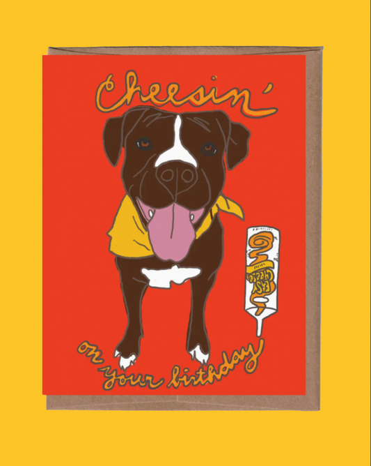 A square greeting card with a brown kraft envelope, featuring a cartoon illustration of a black and white dog wearing a yellow bandana on a bright red background. The card reads 'Cheerin' on your birthday' in yellow script, and a small milk carton graphic says 'Happy Birthday'.