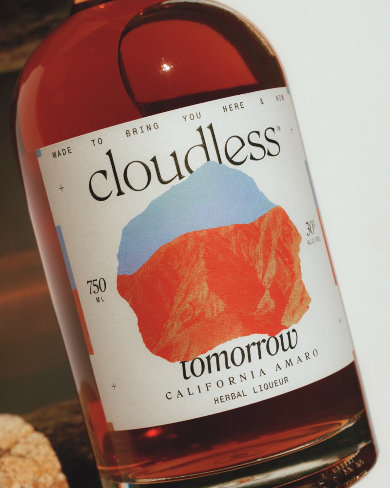 TILTED BOTTLE OF CLOUDLESS TOMORROW CALIFORNIA AMARO HERBAL LIQUEUR ON A WHITE AND BROWN BACKRGOUND