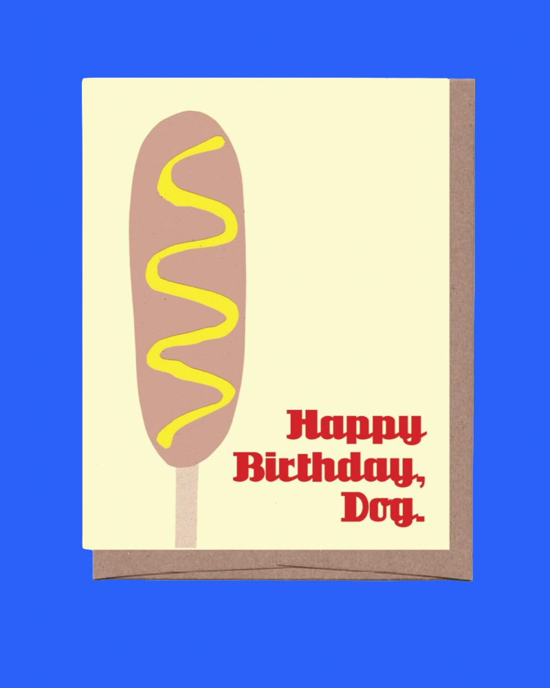 A greeting card featuring a stylized hot dog illustration with mustard drizzle and the text 'Happy Birthday, Dog.' in red, shown with a brown envelope against a solid blue background.