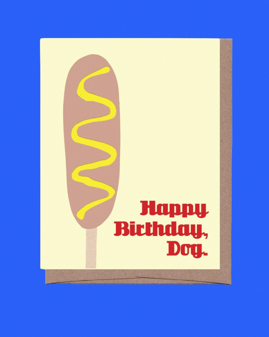 A greeting card featuring a stylized hot dog illustration with mustard drizzle and the text 'Happy Birthday, Dog.' in red, shown with a brown envelope against a solid blue background.