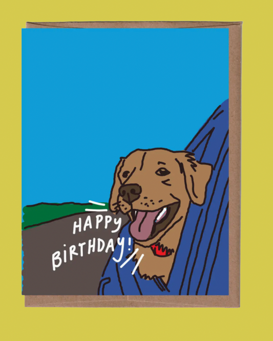 A square greeting card with a brown envelope, featuring a hand-drawn illustration of a brown dog with its tongue out, leaning out of a blue car window. White text on the card reads 'HAPPY BIRTHDAY!'.