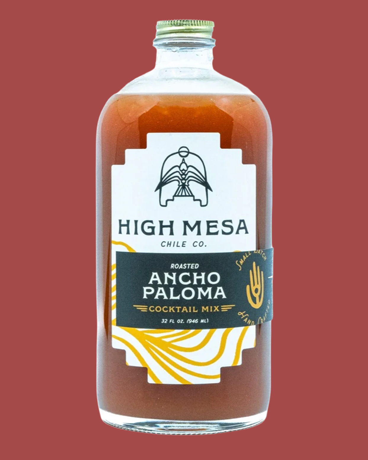 A clear glass bottle of High Mesa Chile Co. Roasted Ancho Paloma Cocktail Mix filled with amber-brown liquid, featuring a white and black label with gold accents, set against a solid muted red background.