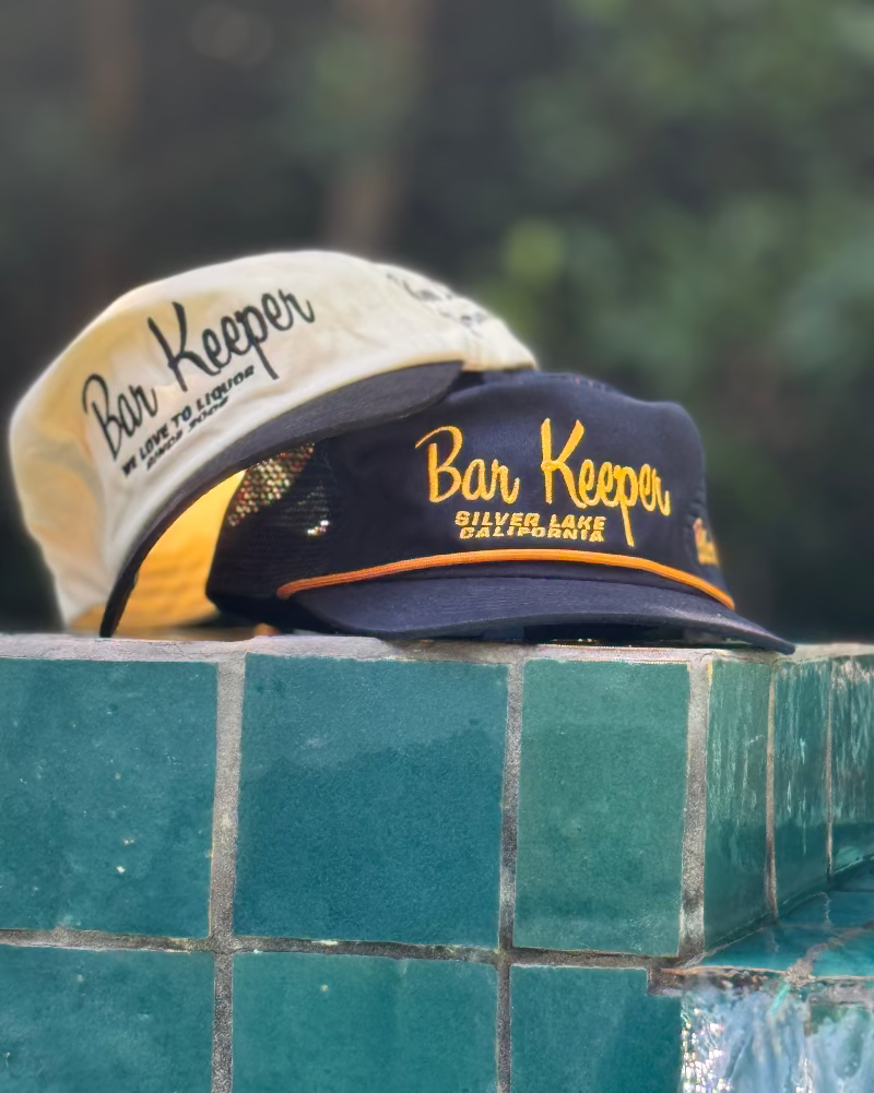 Two 'Bar Keeper' hats on a tiled ledge with a blurred green background