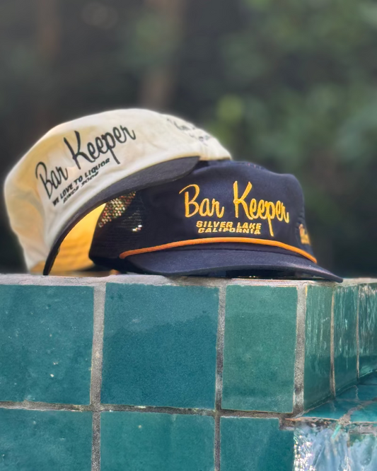 Two 'Bar Keeper' hats on a tiled ledge with a blurred green background