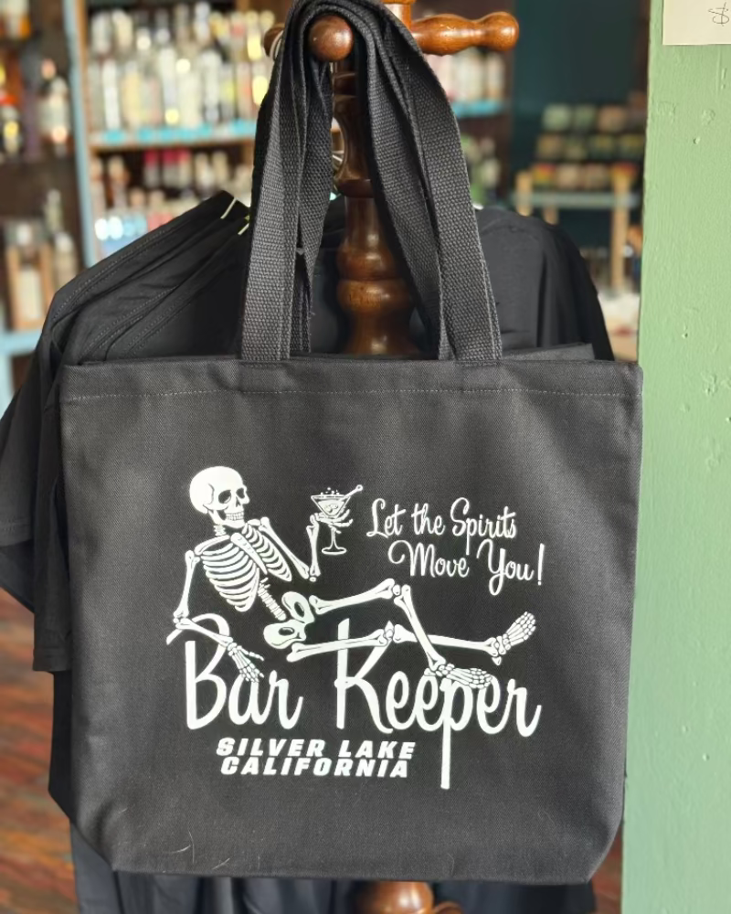 Black tote bag with skeleton design and text, held by a wooden rack in a store setting.