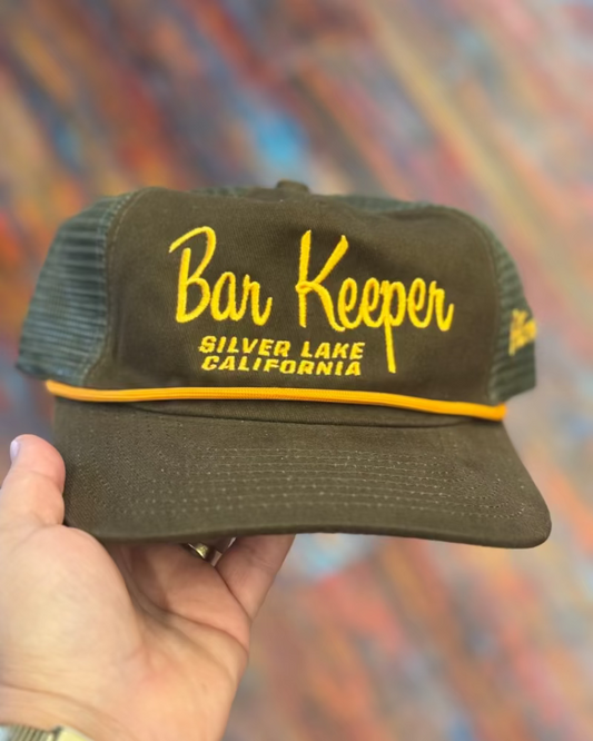 Brown cap with yellow text 'Bar Keeper Silver Lake California' held against a colorful, abstract background.