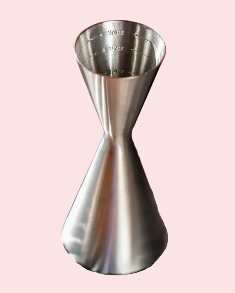 A stainless steel jigger with measurement markings inside, shown on a solid pale pink background.