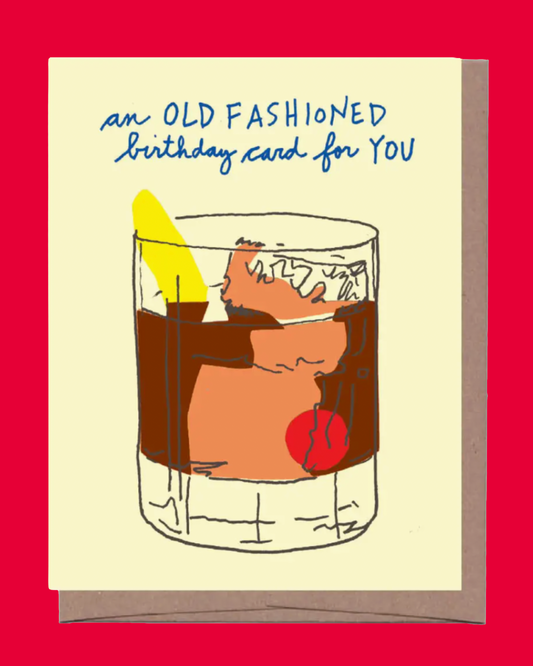 A novelty greeting card with a cream background and blue handwritten text reading 'an OLD FASHIONED birthday card for YOU,' featuring a hand-drawn illustration of a cocktail glass with brown liquid, a yellow lemon peel, and a red cherry. The card is shown against a solid red background with a brown envelope behind it.