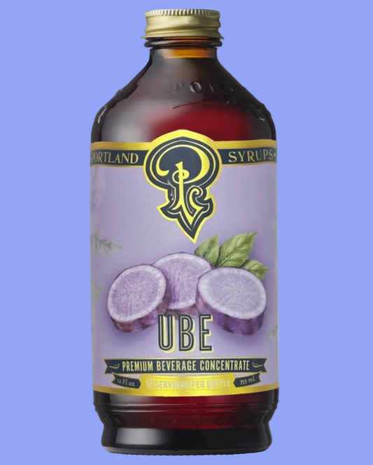 A bottle of Portland Syrup Ube Premium Beverage Concentrate on a clean white background. The bottle is made of amber glass with a gold cap and features a light purple label with illustrations of purple uvafrutto (ube) and green leaves.