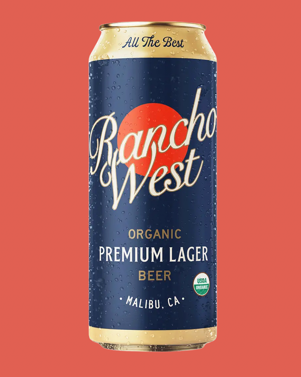 A single can of Rancho West Organic Premium Lager Beer centered on a solid coral-red background. The can is dark blue with a large orange-red sun graphic, white script branding, and gold text accents.