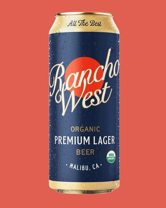 A single can of Rancho West Organic Premium Lager Beer centered on a solid coral-red background. The can is dark blue with a large orange-red sun graphic, white script branding, and gold text accents.