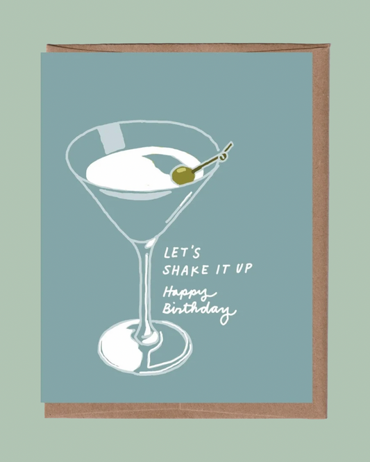 A square greeting card with a dusty blue-green background, featuring a white line art illustration of a martini glass with an olive on a toothpick and the text 'LET'S SHAKE IT UP Happy Birthday'. The card is shown with a brown kraft envelope behind it against a matching sage green background.