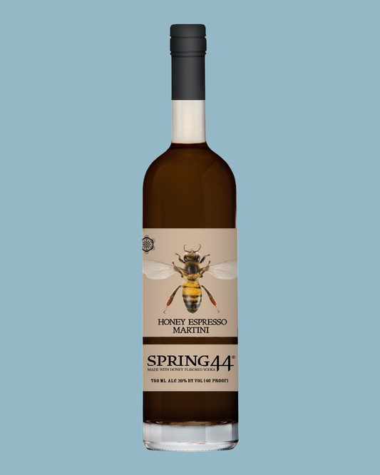 A bottle of Spring44 Honey Espresso Martini liqueur centered on a solid light blue background. The bottle is made of dark glass showing the dark brown liquid inside, with a beige label featuring a detailed illustration of a bee and the product name 'HONEY ESPRESSO MARTINI' and brand 'SPRING44'.