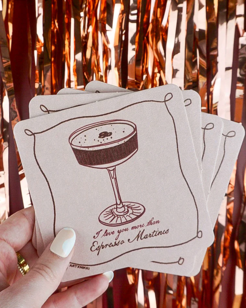 Coasters with a cocktail illustration and text held by a hand against a blurred background