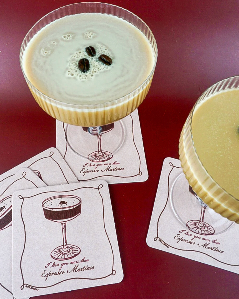 Two cocktails in martini glasses on a red surface with matching coasters.