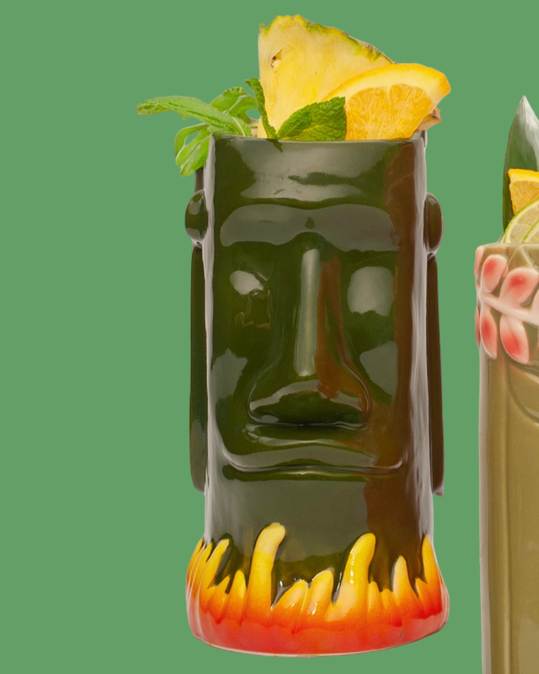 Tiki mug with a face design filled with a cocktail garnished with pineapple and mint leaves on a green background.