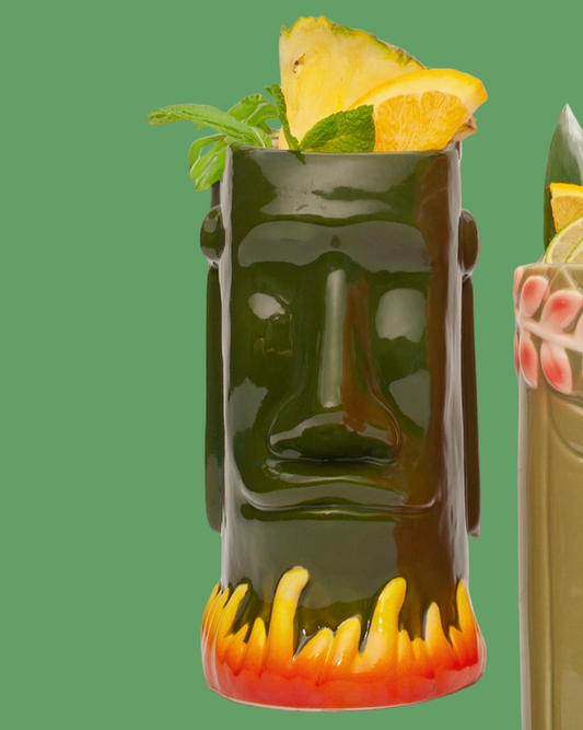 Tiki mug with a face design filled with a cocktail garnished with pineapple and mint leaves on a green background.