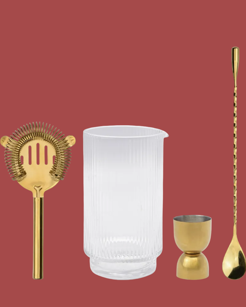 Set of bar tools including a gold strainer, clear glass, gold jigger, and gold spoon on a red background