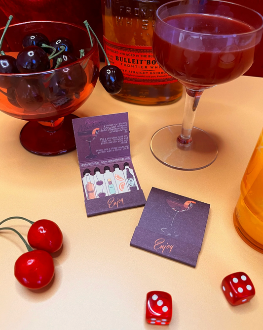 Cocktail setup with a glass of red liquid, bottle, cherries, and dice on a table.