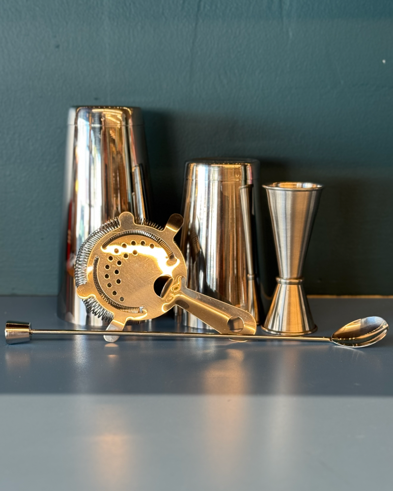 Set of metallic cocktail shakers and tools on a gray surface with a dark green background
