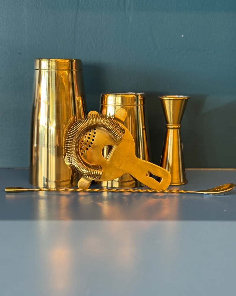 Set of gold cocktail shakers and tools on a gray surface with a blue background