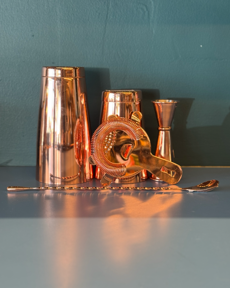 Copper cocktail shaker, jigger, and strainer on a reflective surface with a teal wall background.