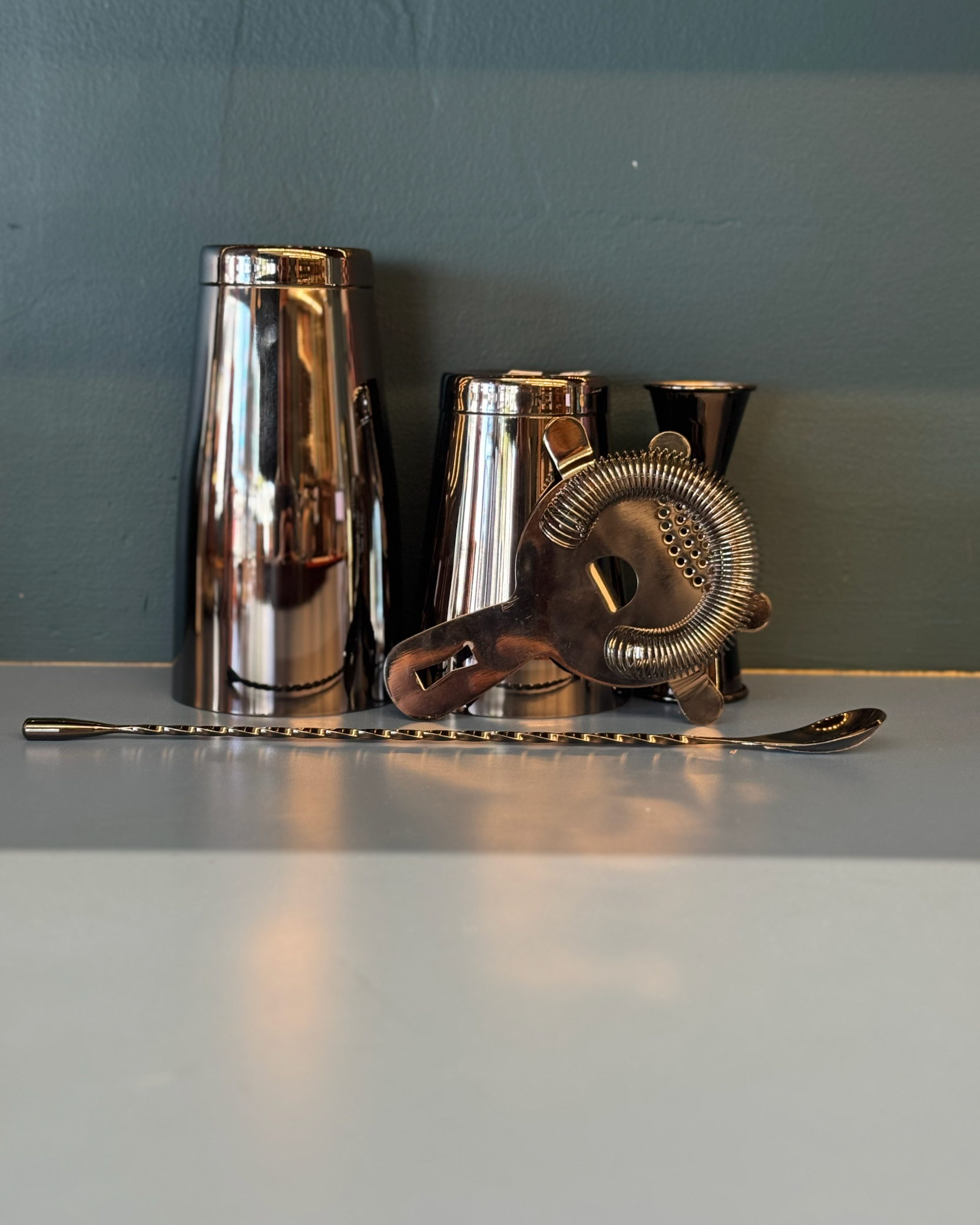 Set of metallic cocktail shakers and a bar spoon on a reflective surface with a gray background