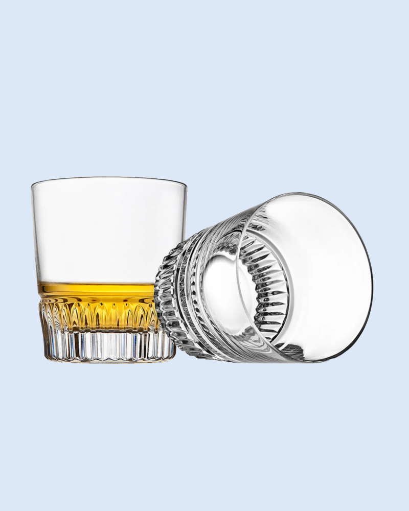 Clear glass with a textured base and rim, containing a small amount of amber liquid on a light blue background.