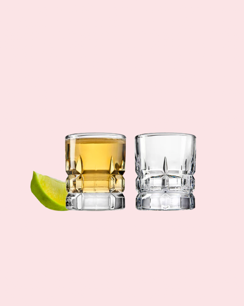 Two glasses with one containing a yellow liquid and a lime wedge on a light pink background
