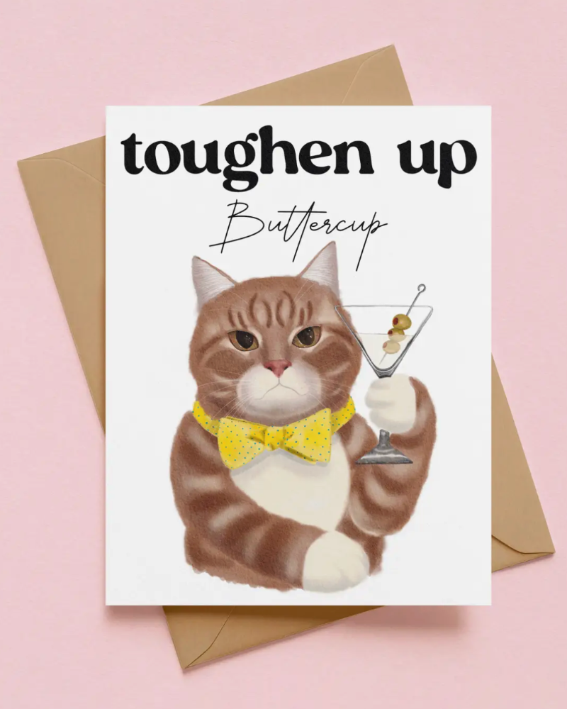 A greeting card featuring a watercolor illustration of a brown and white cat wearing a yellow bow tie and holding a martini glass, with the text 'toughen up Buttercup'. The card is displayed on top of a brown envelope against a solid pink background.