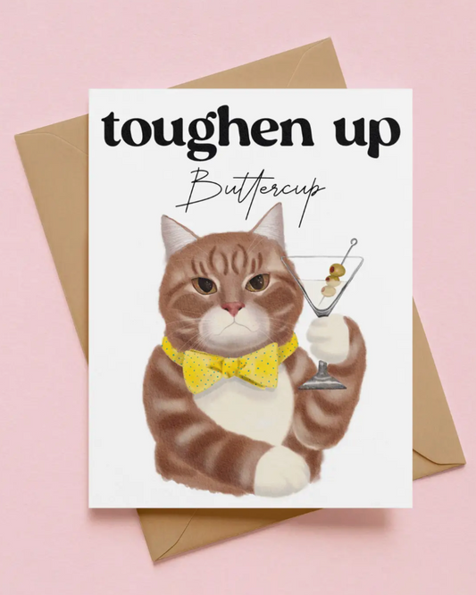 A greeting card featuring a watercolor illustration of a brown and white cat wearing a yellow bow tie and holding a martini glass, with the text 'toughen up Buttercup'. The card is displayed on top of a brown envelope against a solid pink background.