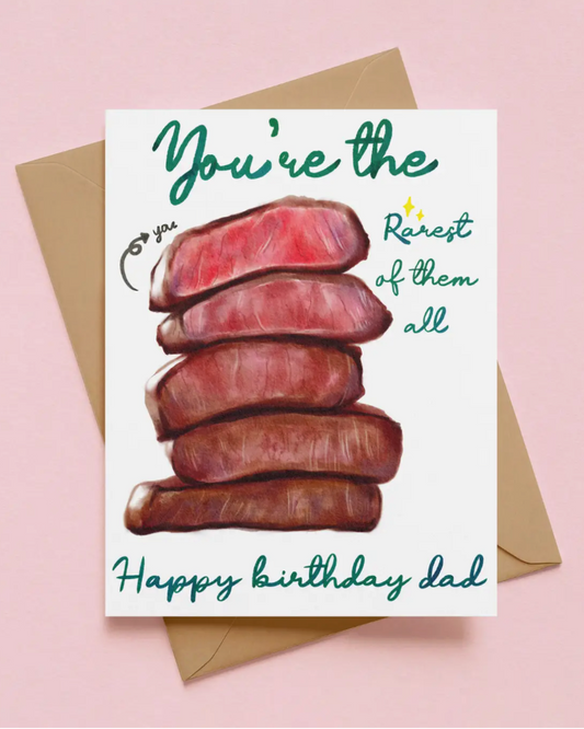 A birthday card with a watercolor illustration of stacked cooked meat and teal script text reading 'You're the Rarest of them all' and 'Happy birthday dad,' shown with a kraft envelope on a pink background.