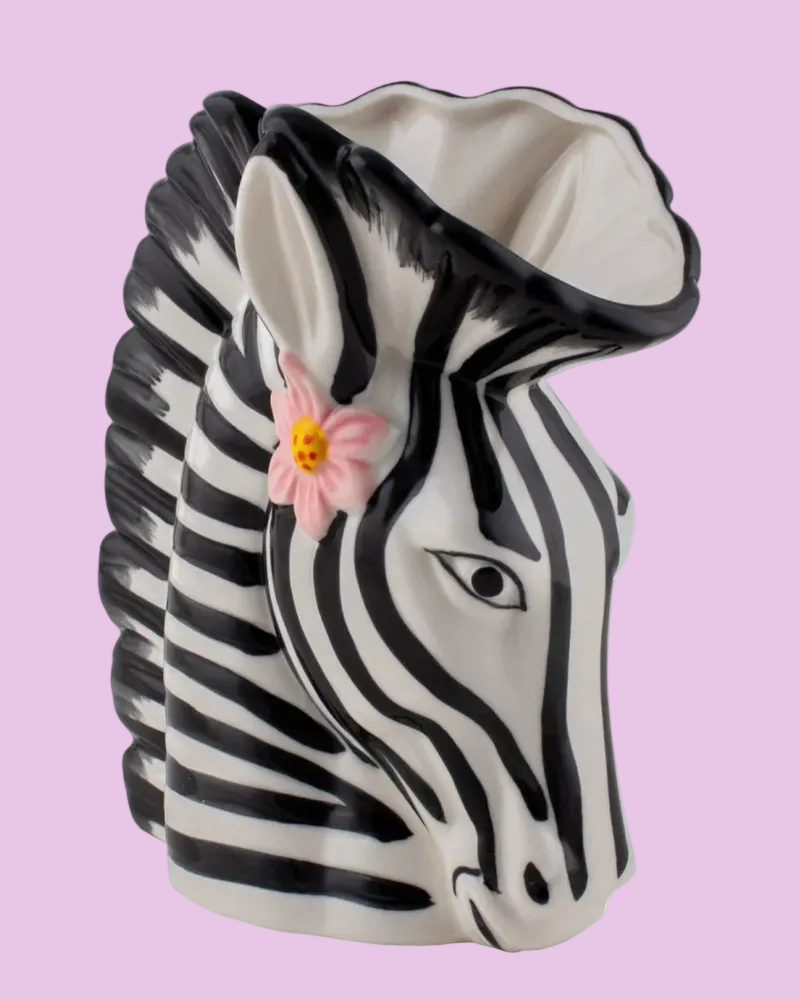 A ceramic zebra head planter with black and white stripes, featuring a small pink flower accent on its side, set against a solid light pink background.